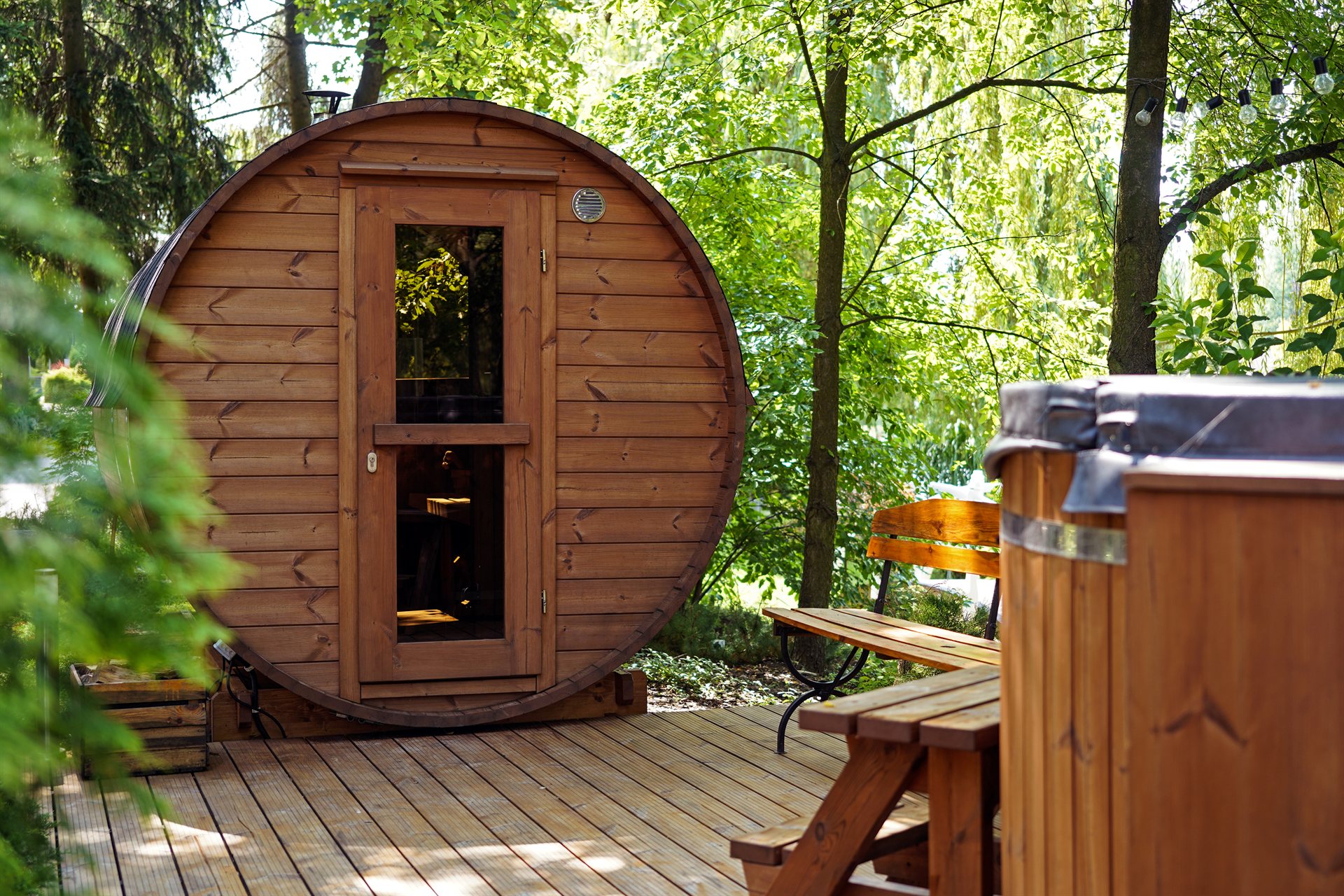 Okrągła drewniana sauna w kształcie beczki na tarasie wśród zielonych drzew, obok ławka i jacuzzi